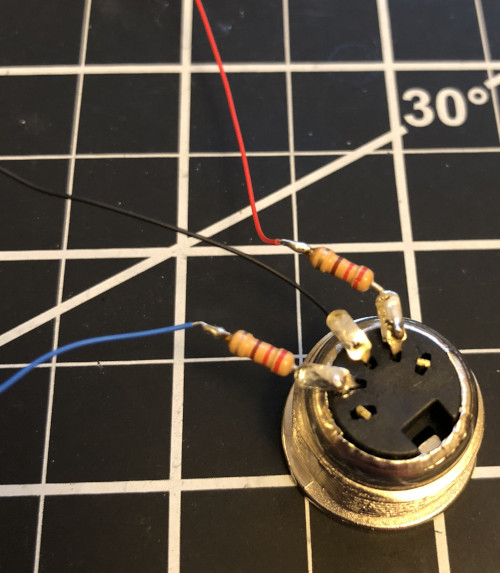 The back of a MIDI connector with the two outer pins clipped and the three inner pins bent up.  Resistors are soldered to the left-most pin and the right-most pin.  The other leads of the resistors are short.  There are wires soldered to the two resistors and to the center pin.
